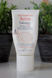 A tube of Avene Mask stood on a white desk with blurred herb plant pots in the background.