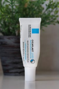 A small white squeeze tube of lip balm standing up right on a white table top. The background is blurred but you can make out a herb plant pot, green herb, grey pot. The lip balm tube has black and bright blue writing: La Roche Posay Cicaplast Leveres