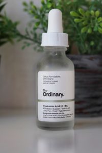 A bottle of serums sitting on a white desk top. Behind the bottles, slightly out of focus are small herbs pots, green herbs in grey pots. The Ordinary hyaluronic acid (clear body, white label, black writing).
