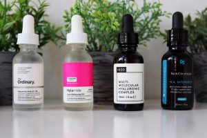 Four bottles of serums sitting on a white desk top. Behind the bottles, slightly out of focus. are three small herbs posts, green herbs in grey pots. The serum bottles from left to right are: The Ordinary hyaluronic acid (clear body, white label, black writing). Hyalmide hyaluronic acid, clear bottle, pink and white label, black writing. NIOD Muli-molecular hyaluronic acid, dark brown bottle, white label, black writing. Skinceuticals H.A Intensifier, Brown bottle, White and bright blue writing.