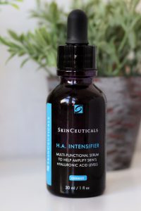 A bottle of serum sitting on a white desk top. Behind the bottles, slightly out of focus are small herbs pots, green herbs in grey pots. SkinCeuticals H.A Intensifier hyaluronic acid (black body, white and bright blue writing).